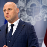 Андрис Кулбергс выступает перед микрофоном на фоне государственного флага.