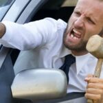 Разгневанный мужчина высовывается из окна автомобиля и указывает рукой, в другой руке у него молоток.