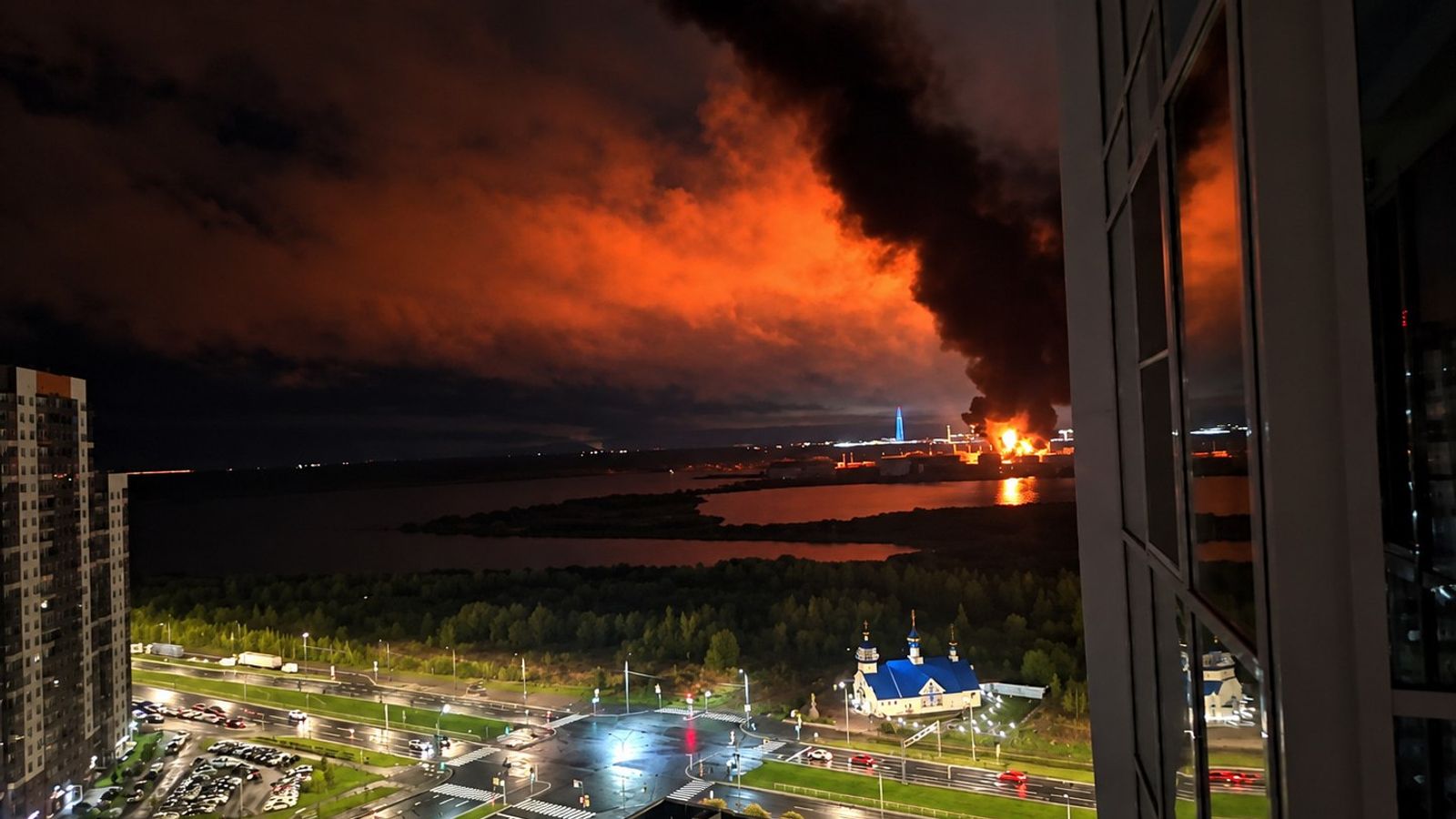 Ночной вид на крупный пожар с густым дымом над городом и водоемом в Санкт-Петербурге.
