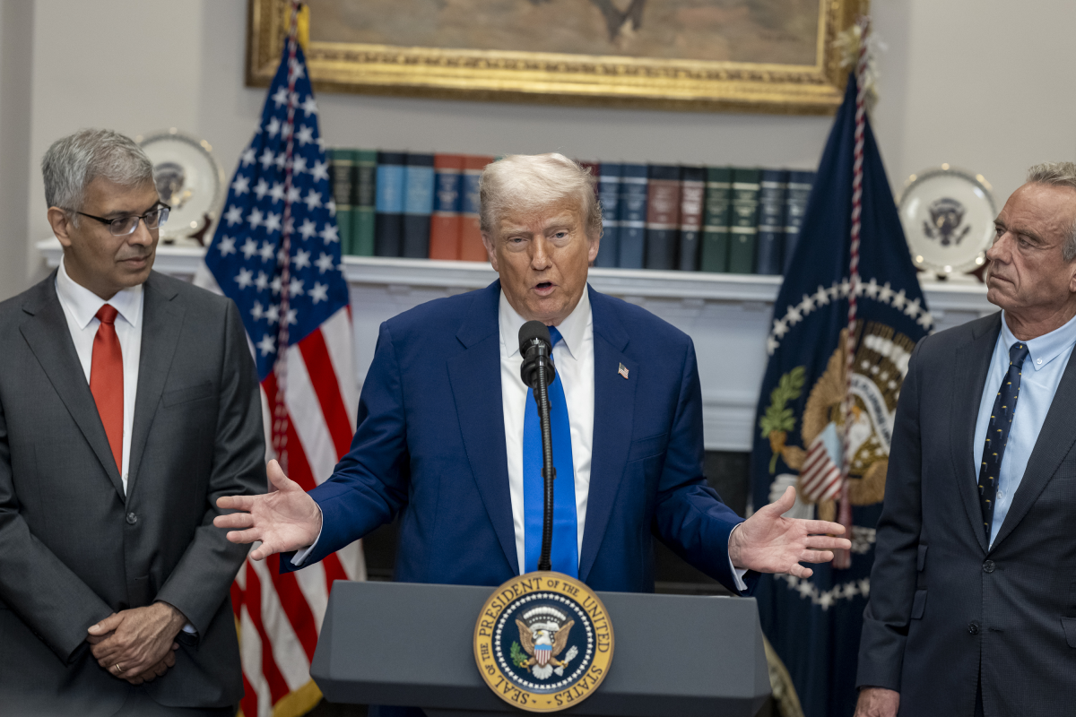 Дональд Трамп выступает у трибуны в официальном помещении, рядом стоят два мужчины в костюмах.