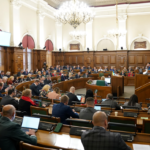 Зал заседаний парламента с депутатами за рабочими местами и трибуной.