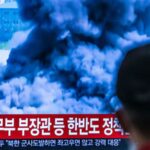 Люди смотрят телевизионный экран с новостным сюжетом и облаком дыма на фоне корейского текста.