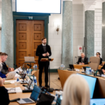 Человек выступает у трибуны на заседании в официальном зале, вокруг сидят участники с ноутбуками и документами.