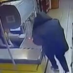 Кадр с камеры наблюдения в продуктовом магазине: человек в темной одежде стоит у кассовой зоны рядом с поврежденным оборудованием.