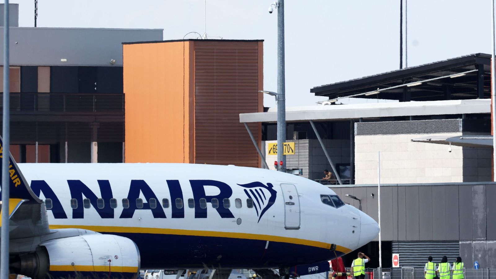 Самолёт Ryanair у терминала аэропорта, на фоне служебных зданий и сотрудников в жилетах.