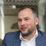 Портрет мужчины в синем клетчатом пиджаке и белой рубашке в помещении.