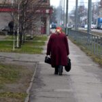 Пожилая женщина идет по тротуару с сумками в руках.