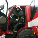 Пожарный стоит в открытой кабине красной пожарной машины.