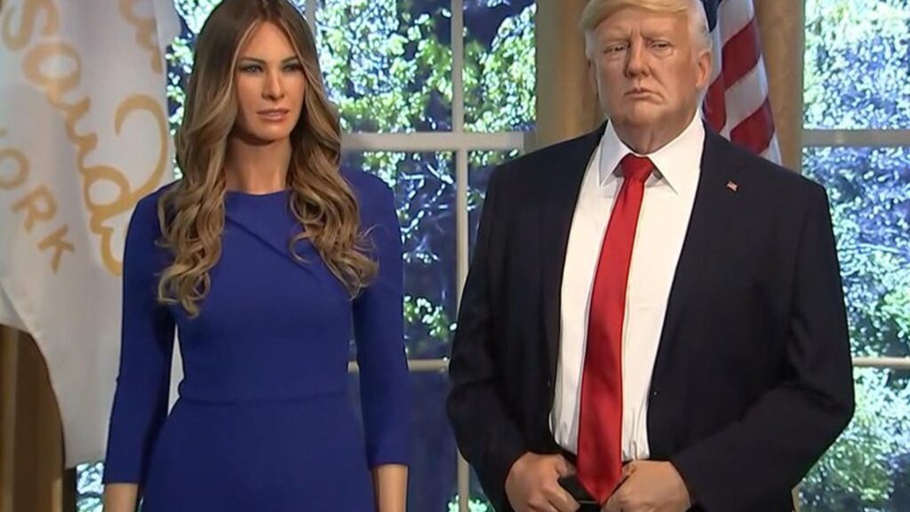 Мелания Трамп и Дональд Трамп стоят в помещении перед окном и американским флагом.