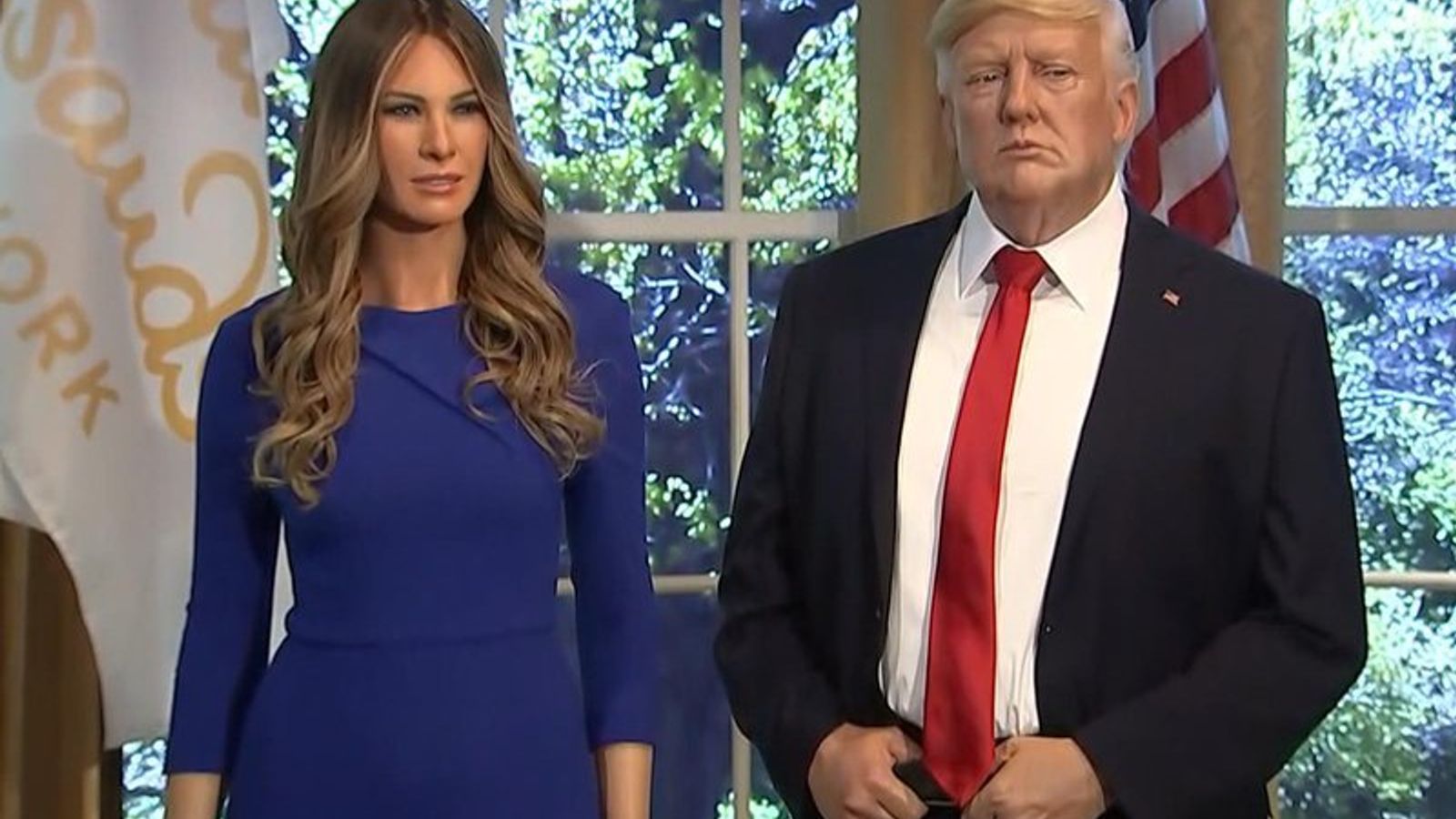 Мелания Трамп и Дональд Трамп стоят в помещении перед окном и американским флагом.