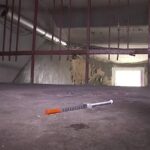 Шприц лежит на полу в полутёмном помещении с повреждёнными стенами и металлическими конструкциями.
