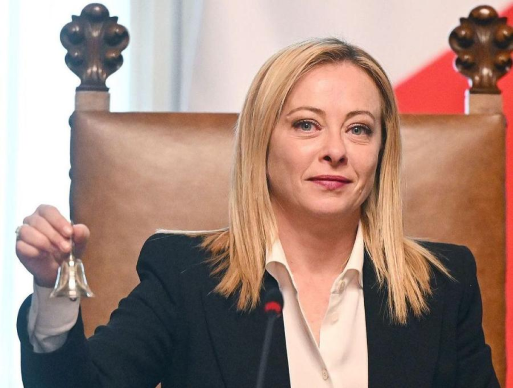 Женщина в деловом костюме сидит в кресле и держит маленький колокольчик.