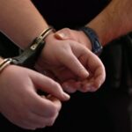 Руки человека в наручниках, удерживаемые сотрудником полиции.