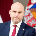 Андрис Кулбергс в костюме и красном галстуке на фоне латвийского флага и герба.
