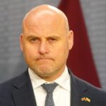 Портрет лысого мужчины в костюме и галстуке с латвийским значком на лацкане.
