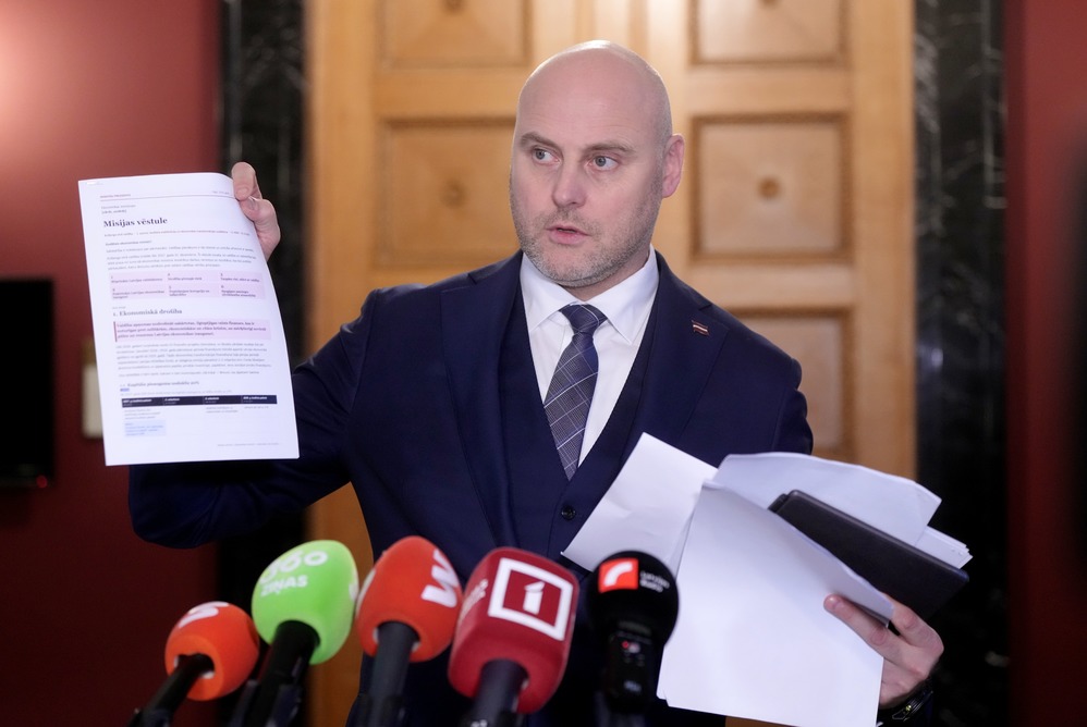 Мужчина в костюме показывает листы документов перед микрофонами на пресс-конференции.