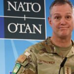 Алексус Гринкевич в военной форме на фоне логотипа НАТО.