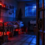 Интерьер тюремной камеры с кроватью, решётками на окне и красно-синим освещением.