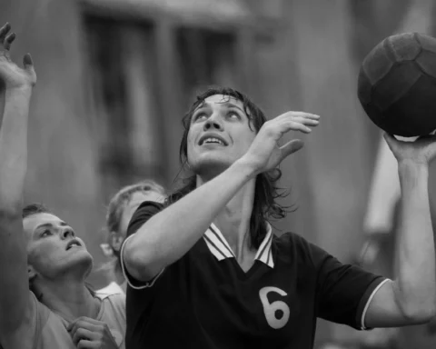 Черно-белый кадр: женщина в спортивной форме с номером 6 держит мяч и готовится к броску, рядом другие игроки тянутся вверх.