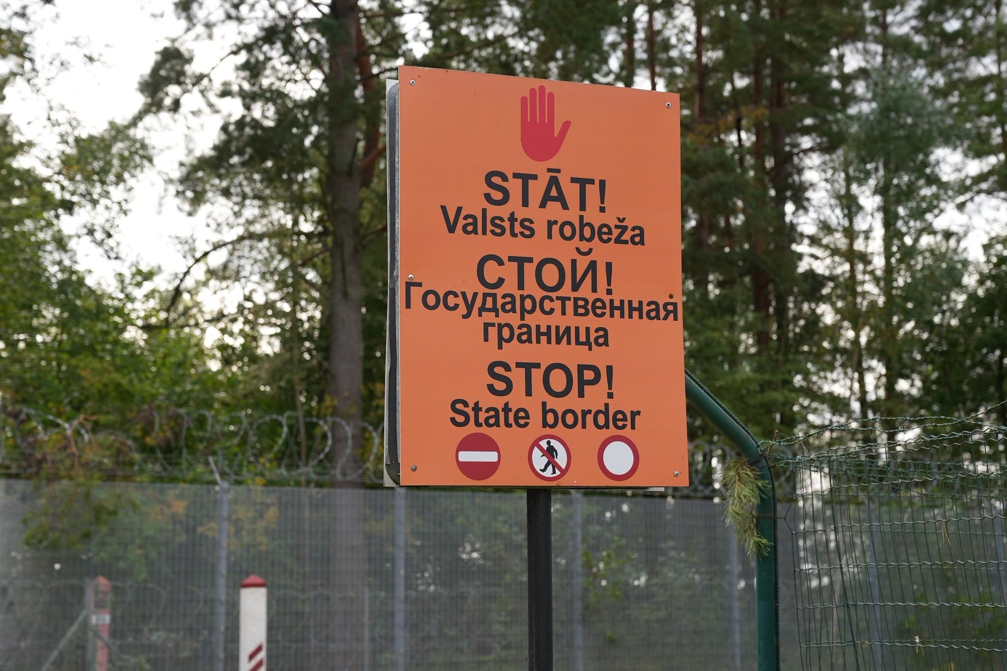 Orange warning sign at the Latvian state border near a fenced wooded area.