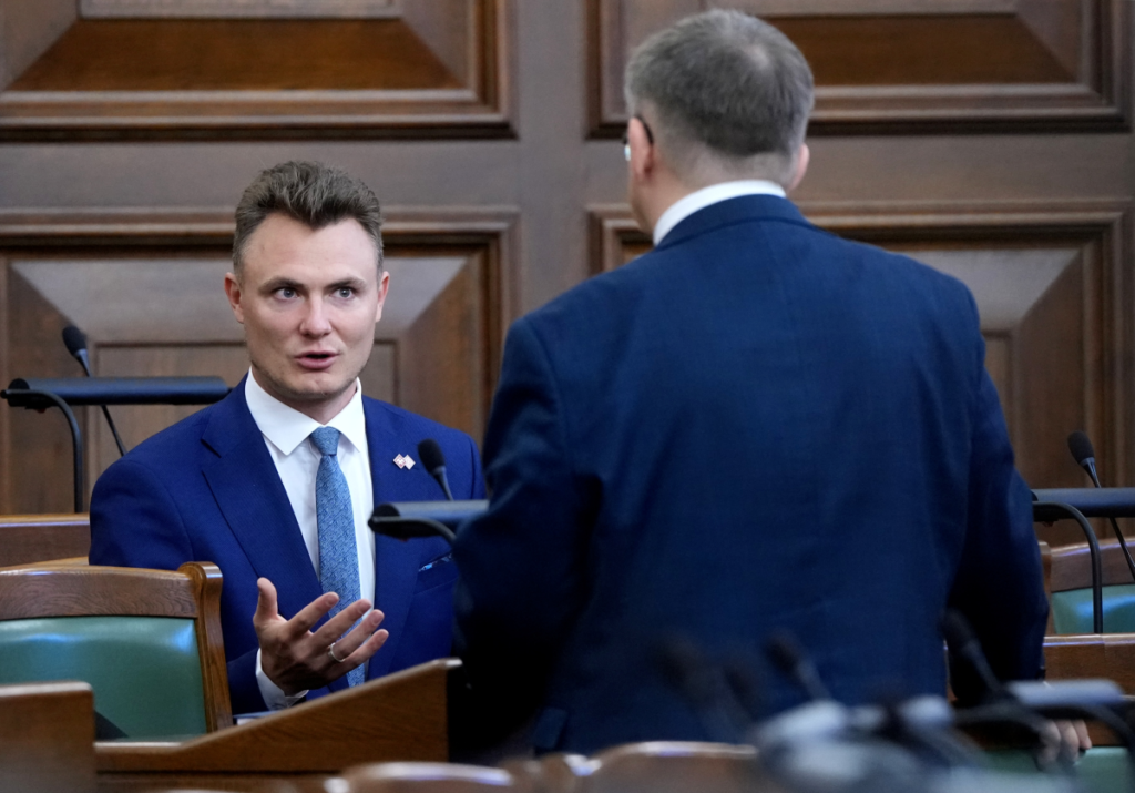 Депутаты в зале заседаний парламента Латвии, один из них выступает, обращаясь к другому.