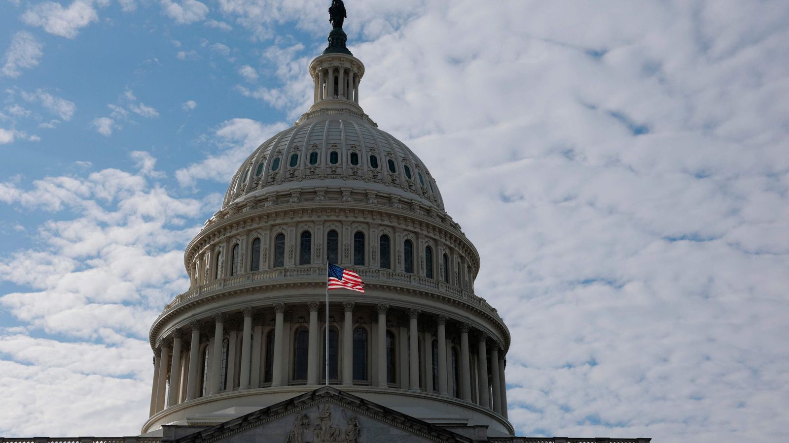Купол здания Капитолия США на фоне облачного неба, на фасаде виден американский флаг.