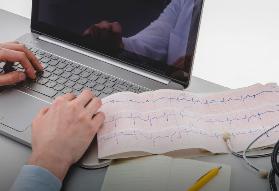Hands typing on a laptop beside an ECG printout and stethoscope on a desk.