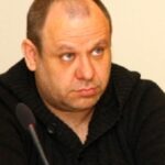Александр Жгун сидит за микрофоном на фоне светлой стены.