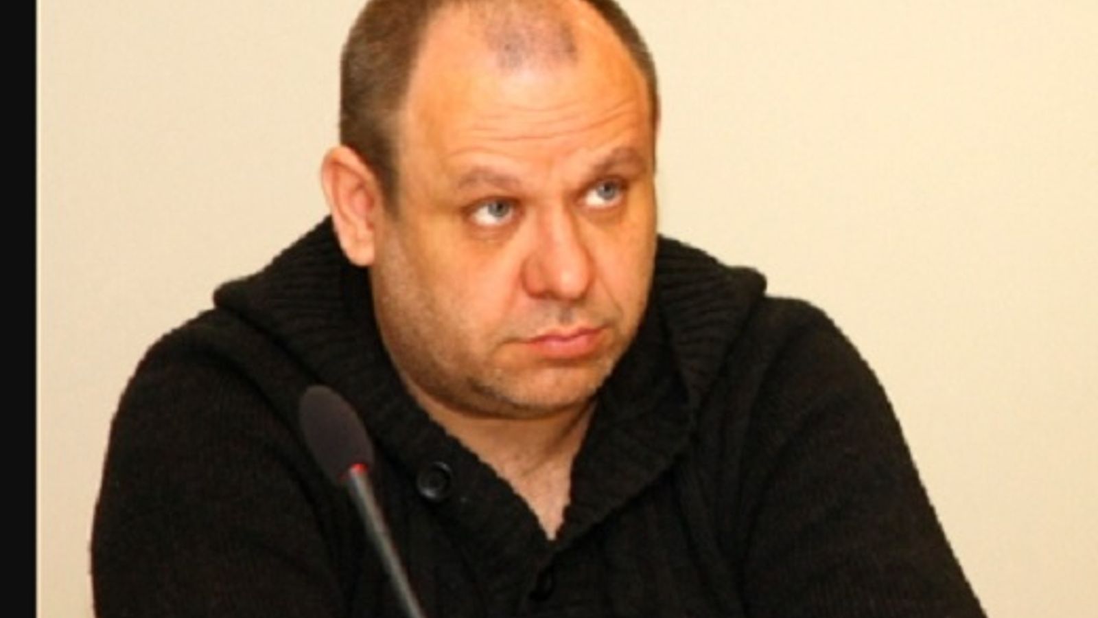 Александр Жгун сидит за микрофоном на фоне светлой стены.