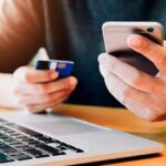 Человек держит банковскую карту и смартфон рядом с ноутбуком.