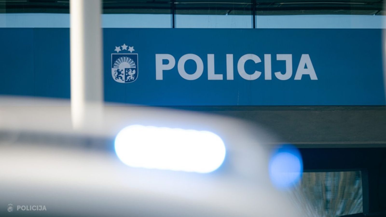 Здание или вход полиции с крупной надписью «POLICIJA» и размытыми синими огнями автомобиля на переднем плане.