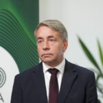Улдис Аугулис в костюме на фоне зелёного фирменного стенда.