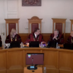Судьи Конституционного суда Латвии сидят в зале заседаний за длинным столом.