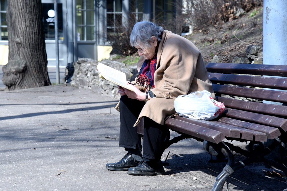 Пожилая женщина сидит на скамейке в парке и читает книгу.