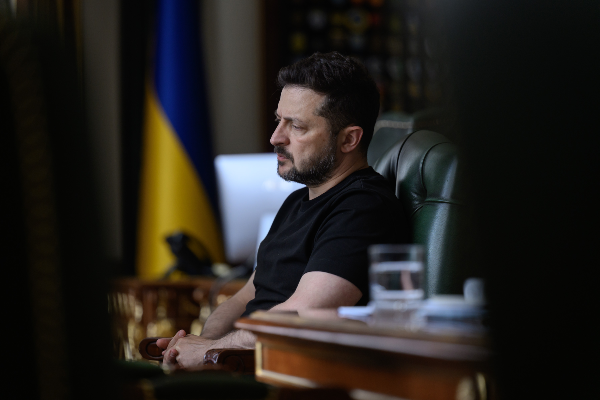Владимир Зеленский сидит в кресле в офисе на фоне украинского флага.
